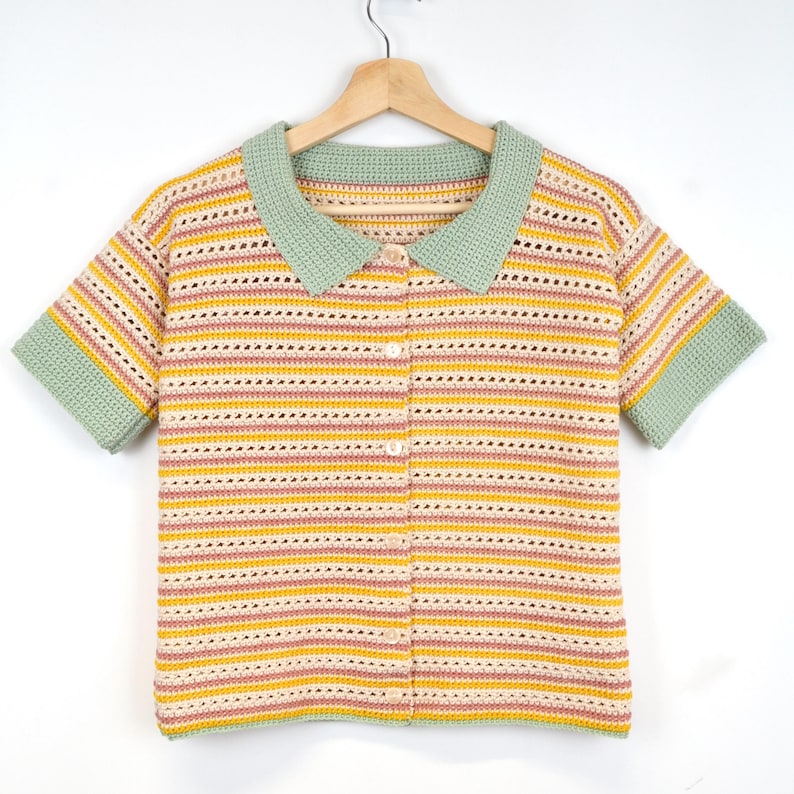 Puede incluir: Un top de crochet de manga corta, a rayas, con cuello verde y cierre de bot&oacute;n. El top est&aacute; hecho con hilo de color rosa claro, amarillo y crema.