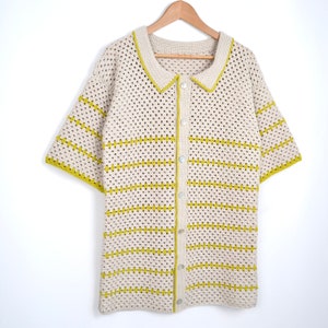 Puede incluir: Una camisa de crochet color crema, de manga corta, con botones en la parte delantera y cuello. La camisa presenta rayas horizontales y ribetes en un color amarillo verdoso brillante. La camisa est&aacute; colgada en una percha de madera sobre un fondo blanco.