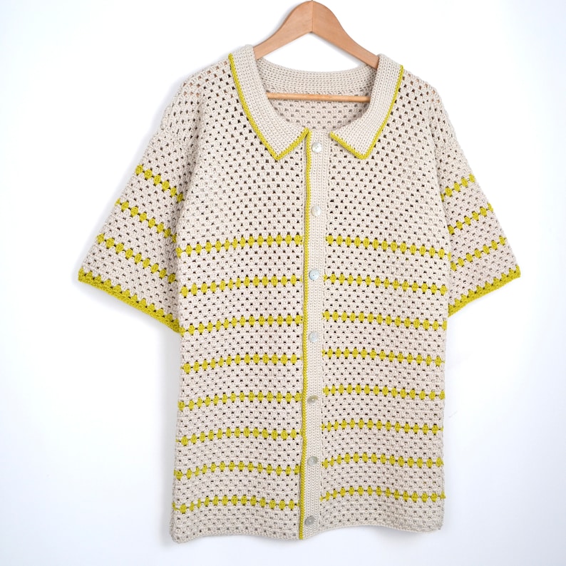 Puede incluir: Una camisa de crochet color crema, de manga corta, con botones en la parte delantera y cuello. La camisa presenta rayas horizontales y ribetes en un color amarillo verdoso brillante. La camisa est&aacute; colgada en una percha de madera sobre un fondo blanco.