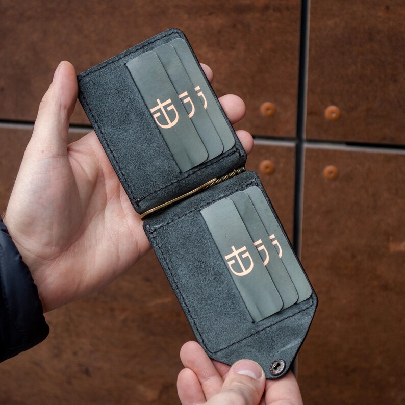 May include: A black leather money clip wallet featuring two interior sections, each with three card slots. The card slots are accented with a light green leather trim and a copper-colored logo symbol. A gold-toned metal money clip is attached.