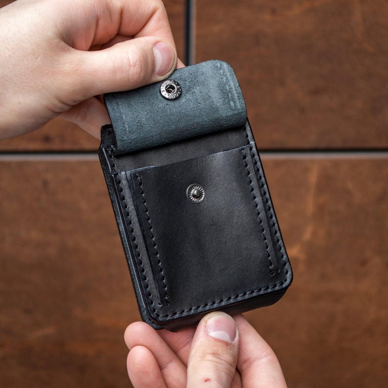May include: A black leather pouch featuring a rectangular shape with visible stitching along the edges. The pouch includes a flap closure secured by a silver-toned metal snap button, with an additional snap button located on the front pocket surface.