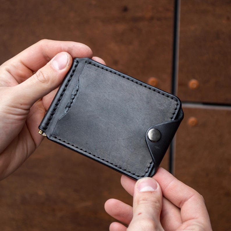 May include: A black leather money clip wallet featuring visible stitching along the edges, a curved card slot opening on the front, and a single metallic snap button closure on the side to secure the folded leather flap over the main compartment.
