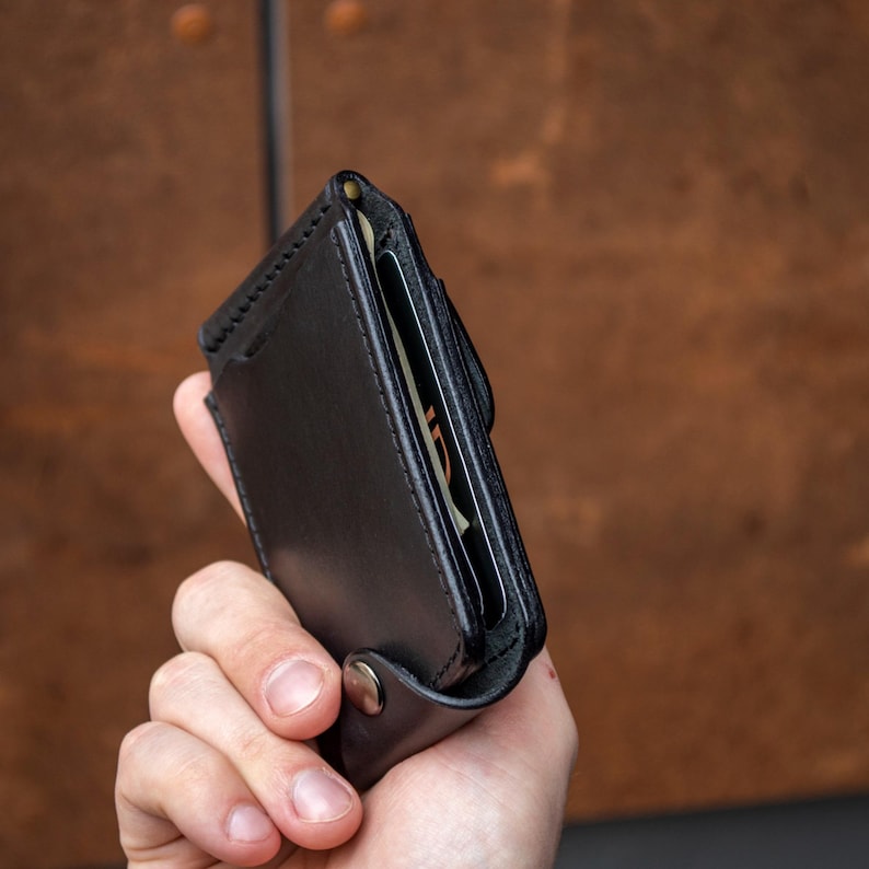 May include: A black leather card holder wallet featuring visible stitching along the edges, a metallic snap button closure at the bottom, and an open top slot containing a card with visible text, constructed with a slim profile and smooth finish.