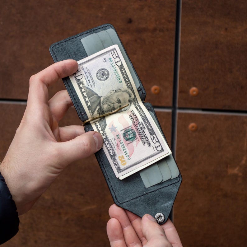 May include: A black leather money clip wallet featuring a central gold-toned metal clip holding a fifty-dollar bill. The wallet includes multiple interior card slots with grey accents and a single silver-toned snap button closure at the bottom edge.