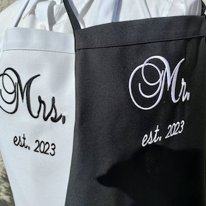 May include: Two aprons, one white and one black, are displayed. The white apron has black embroidered text reading "Mrs. est. 2023." The black apron has white embroidered text reading "Mr. est. 2023."