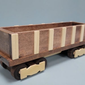 May include: A wooden toy train car with a brown body and white trim. The car has four wheels and a hook on the front.