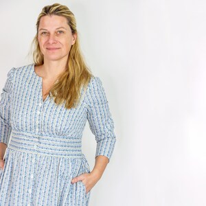 May include: A light blue dress with a vertical striped pattern and small floral accents. The dress features a V-neckline, button-down front, and a fitted waist. The sleeves are three-quarter length, and the dress has pockets.