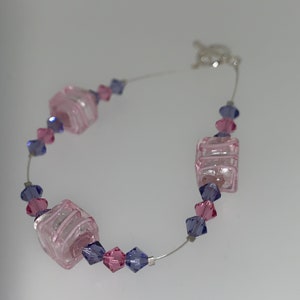 May include: A delicate bracelet with pink and purple glass beads. The bracelet features square-shaped beads with a layered design and smaller, faceted beads.