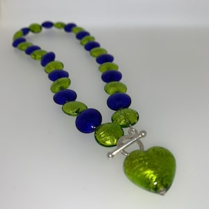 May include: A green and blue glass bead necklace with a green glass heart pendant. The necklace has a silver toggle clasp.