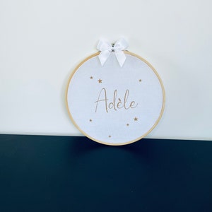 Peut inclure: Un cercle de broderie blanc avec le nom "Adèle" brodé en fil doré et entouré d'étoiles dorées. Le cercle est suspendu à un mur blanc avec un mur noir en dessous.
