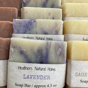May include: Close-up of several handmade soap bars. The bars are various colors, including lavender and sage. The lavender soap has a marbled purple and white design. Each bar is wrapped with a label that reads "Heathers Natural Home".