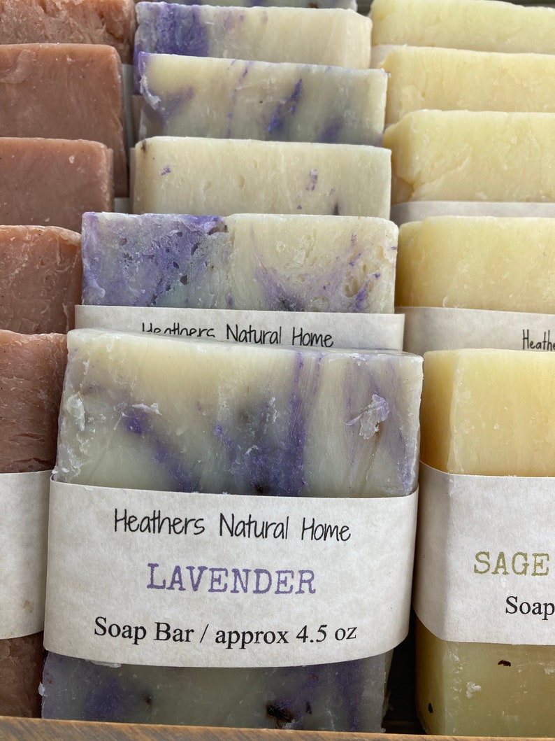 May include: Close-up of several handmade soap bars. The bars are various colors, including lavender and sage. The lavender soap has a marbled purple and white design. Each bar is wrapped with a label that reads "Heathers Natural Home".