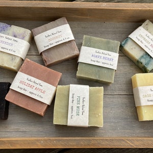 May include: Assortment of handmade soap bars in a wooden tray. Scents include Lavender, Coconut, Holiday Apple, Agave Berry, Pine Musk, Cypress Berry, Sage & Lemongrass. Each bar is wrapped with a label indicating the scent and weight, approximately 4.5 oz.