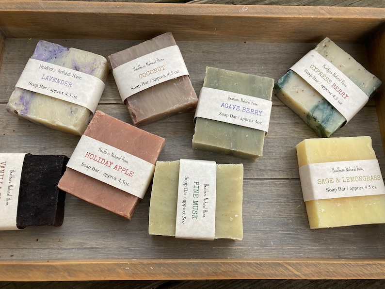 May include: Assortment of handmade soap bars in a wooden tray. Scents include Lavender, Coconut, Holiday Apple, Agave Berry, Pine Musk, Cypress Berry, Sage & Lemongrass. Each bar is wrapped with a label indicating the scent and weight, approximately 4.5 oz.