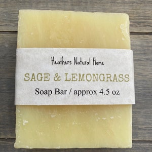 May include: A rectangular bar of soap with a light yellow hue, wrapped with a paper label. The label reads "Heathers Natural Home" and "SAGE & LEMONGRASS Soap Bar / approx 4.5 oz". The soap is presented on a wooden surface.