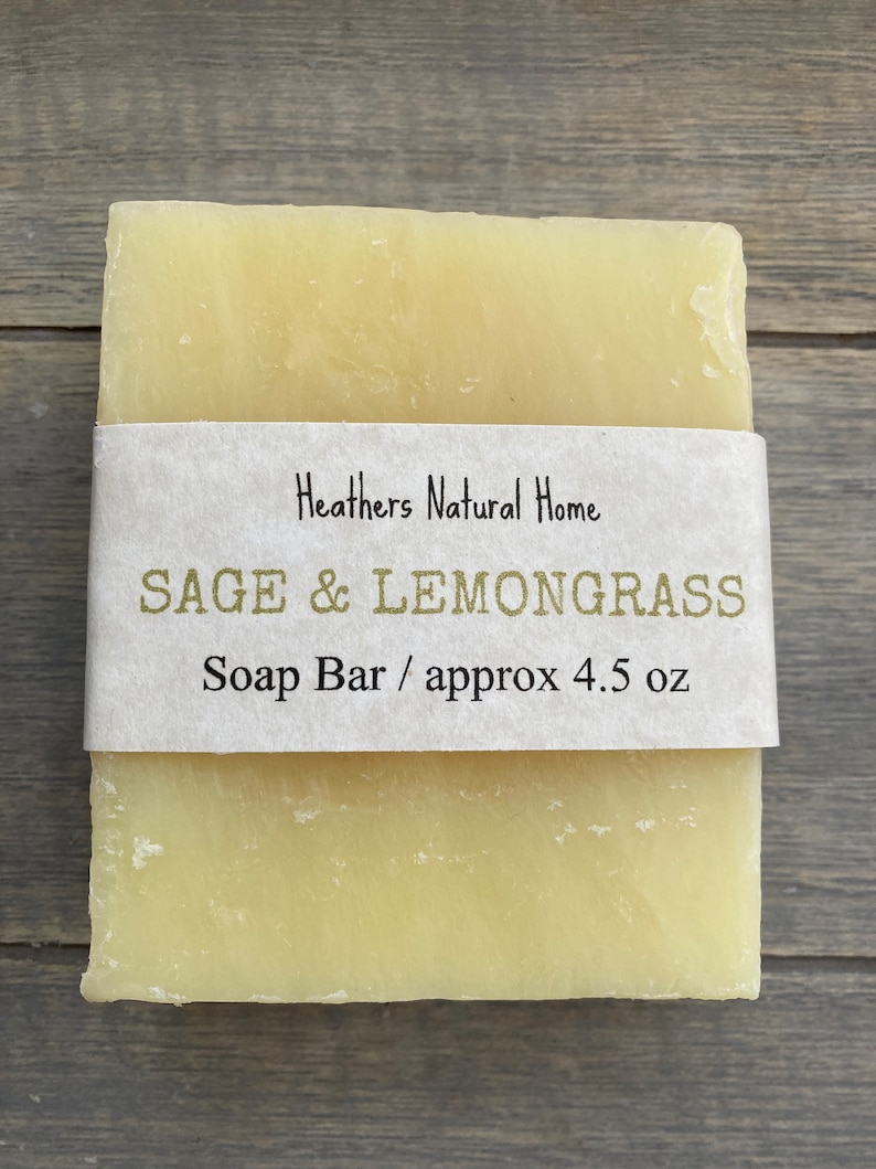 May include: A rectangular bar of soap with a light yellow hue, wrapped with a paper label. The label reads "Heathers Natural Home" and "SAGE & LEMONGRASS Soap Bar / approx 4.5 oz". The soap is presented on a wooden surface.
