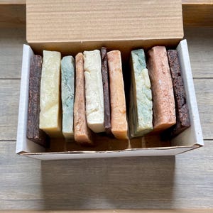 May include: A box of assorted handmade soaps in various colors and textures. The soaps are rectangular and feature shades of brown, beige, green, and orange. The box is made of cardboard and sits on a wooden surface.