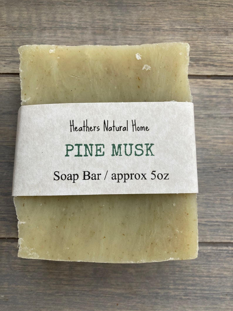 May include: A rectangular bar of soap with a light green hue, wrapped with a white paper band. The band features the text "Heathers Natural Home," "PINE MUSK," and "Soap Bar / approx 5oz."
