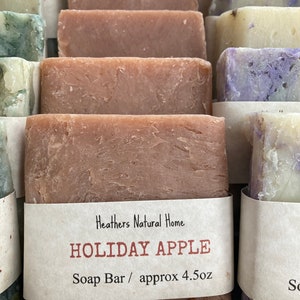 May include: Close-up of several rectangular soap bars in various colors, including shades of brown, green, and purple. One bar is labeled "Holiday Apple" with the text "Soap Bar / approx 4.5oz". The soaps appear to be handmade.