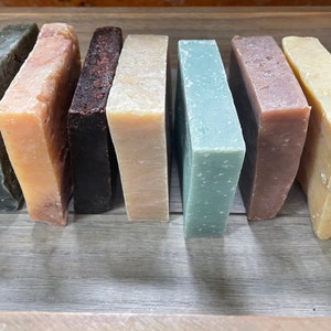 May include: Seven rectangular bars of soap arranged in a row. The colors include dark gray, light peach, dark brown, beige, light sage green, mauve, and pale yellow. Each bar has a solid, block-like shape with a smooth, matte finish and flat edges.