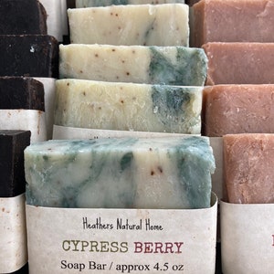 May include: Close-up of several handmade soap bars. The bars are various colors, including white, green, and brown. One bar is labeled "Cypress Berry" and is approximately 4.5 oz. The soaps are arranged in rows, suggesting a display.