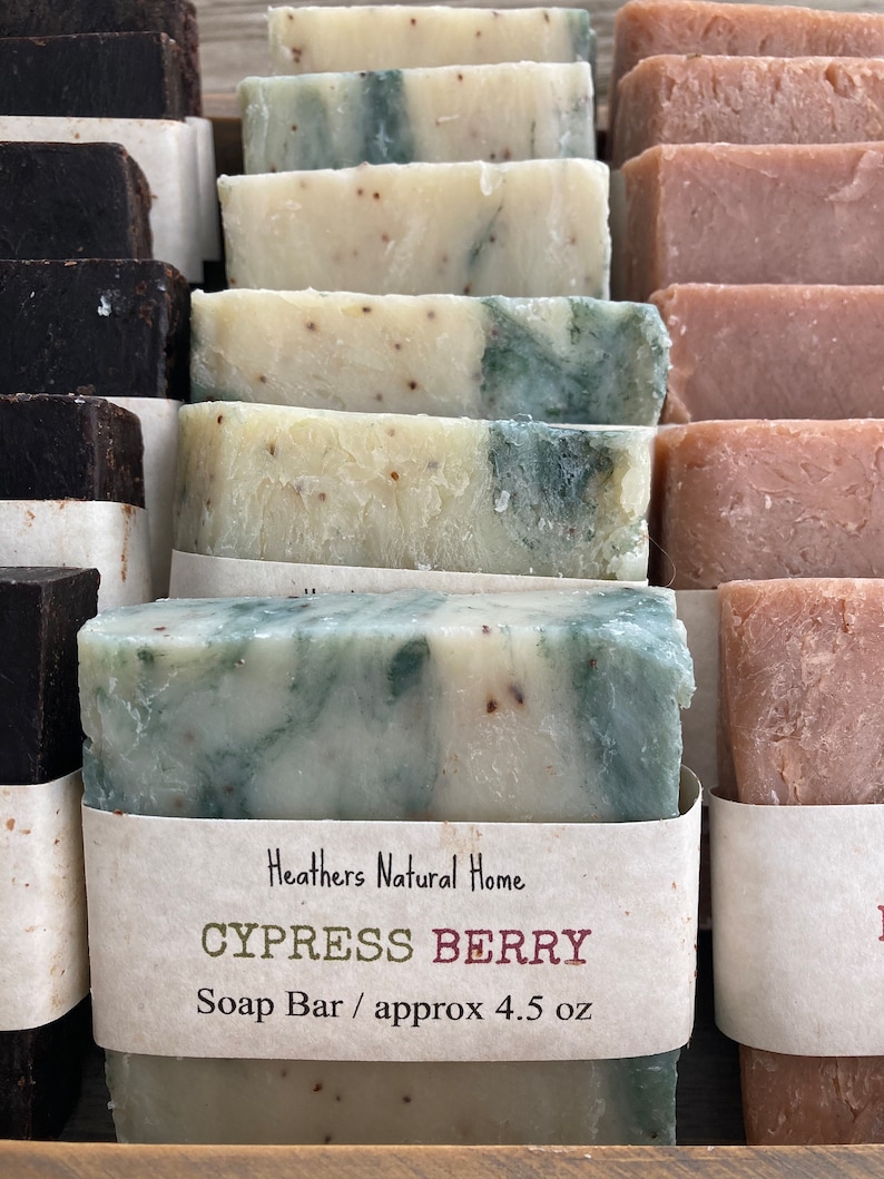 May include: Close-up of several handmade soap bars. The bars are various colors, including white, green, and brown. One bar is labeled "Cypress Berry" and is approximately 4.5 oz. The soaps are arranged in rows, suggesting a display.
