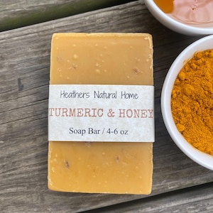 Puede incluir: Una pastilla de jabón rectangular de cúrcuma y miel con una etiqueta blanca que dice "Heathers Natural Home TURMERIC & HONEY Soap Bar". Dos pequeños cuencos blancos, uno con miel y otro con cúrcuma en polvo, se encuentran en el fondo.