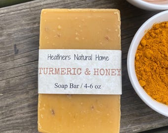 May include: A light orange turmeric and honey soap bar with a white label reading "Heathers Natural Home TURMERIC & HONEY Soap Bar / 113-170 g." Two small white bowls, one with honey and one with turmeric powder, are in the background. The soap is rectangular.
