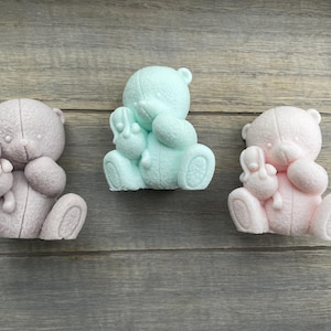May include: Three teddy bear figurines, each holding a smaller bunny figurine. The teddy bears are in different colors: light blue, light pink, and light gray.