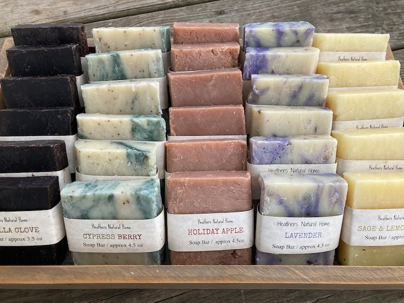 May include: A wooden tray displays various handmade soap bars. The soaps are rectangular and wrapped with labels. Scents include Vanilla Clove, Cypress Berry, Holiday Apple, Lavender, and Sage & Lemon. Each bar weighs approximately 3.5 to 4.5 ounces.