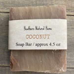 May include: A rectangular bar of soap with a light brown color and a paper label wrapped around it. The label reads "Heathers Natural Home COCONUT Soap Bar / approx 4.5 oz."