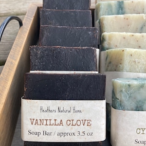 May include: A wooden tray displays several dark brown Vanilla Clove soap bars, each wrapped with a label. The label reads "Vanilla Clove Soap Bar / approx 3.5 oz". Beside them are lighter green and white soap bars.