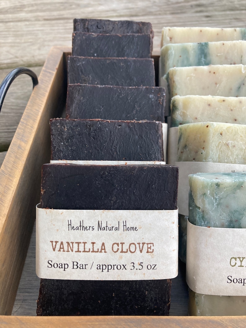May include: A wooden tray displays several dark brown Vanilla Clove soap bars, each wrapped with a label. The label reads "Vanilla Clove Soap Bar / approx 3.5 oz". Beside them are lighter green and white soap bars.
