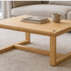 Puede incluir: Una mesa de centro rectangular de madera clara con una superficie lisa y un marco robusto que cuenta con cuatro patas conectadas por soportes de base horizontales. Dos libros apilados y una taza de cerámica descansan sobre la superficie.