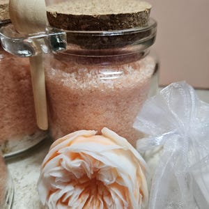 Puede incluir: Un tarro de vidrio con tapa de corcho lleno de sales de baño rosas. Una cuchara de madera está unida al tarro. Una rosa blanca y una bolsa de organza blanca están en el fondo.