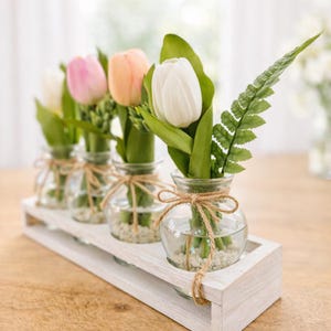 Può includere: Un vassoio decorativo in legno bianco contiene cinque piccoli vasi di vetro, ognuno con un tulipano e del verde. I tulipani sono bianchi, rosa e pesca. I vasi sono legati con spago.