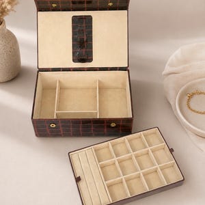 May include: A dark brown, crocodile-textured jewelry box with a hinged lid and gold-tone snap closures. The interior is lined in cream-colored fabric, featuring a removable tray with multiple small square compartments and ring slots.