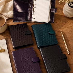 Puede incluir: Una colección de cinco organizadores personales encuadernados en cuero de varios colores, incluyendo morado intenso, verde azulado oscuro y negro. Un organizador está abierto, revelando un mecanismo de anillas metálicas y páginas con líneas con el texto ADDRESS.