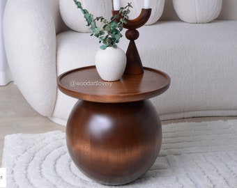May include: A round, dark brown wooden side table with a spherical base and a circular top. A white vase with greenery and a wooden candle holder with two white candles sit on the table. The table is in front of a white sofa.