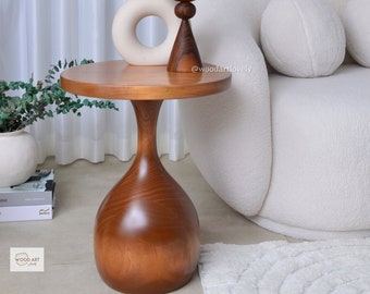 May include: A wooden side table with a round top and a unique, hourglass-shaped base. The table is made of dark wood and has a natural, polished finish. The table is sitting on a white rug in a living room setting.