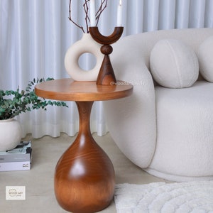 Puede incluir: Una mesa auxiliar de madera con una tapa redonda y una base única en forma de reloj de arena. La mesa está hecha de madera oscura y tiene un acabado natural y pulido. La mesa está sentada sobre una alfombra blanca en un ambiente de sala de estar.