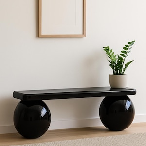 Puede incluir: Un banco negro con una tapa de madera y dos grandes esferas negras para las patas. El banco está frente a una pared blanca con un cuadro enmarcado encima. Una pequeña planta en maceta está a la derecha del banco.