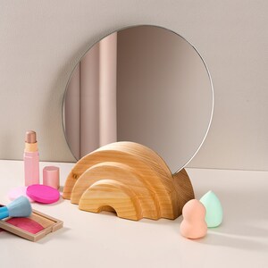 May include: A wooden vanity mirror stand with a round mirror. The stand is shaped like an arch and has a natural wood finish. The mirror is positioned on top of the stand and is slightly angled.
