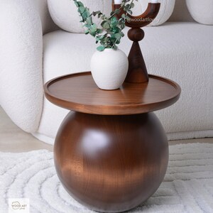 Puede incluir: Una mesa auxiliar moderna de madera marr&oacute;n oscuro con una tapa redonda y una base esf&eacute;rica. La mesa exhibe un jarr&oacute;n blanco con follaje verde y un candelabro de madera. El dise&ntilde;o es minimalista y elegante, con un di&aacute;metro de aproximadamente 60 cm.