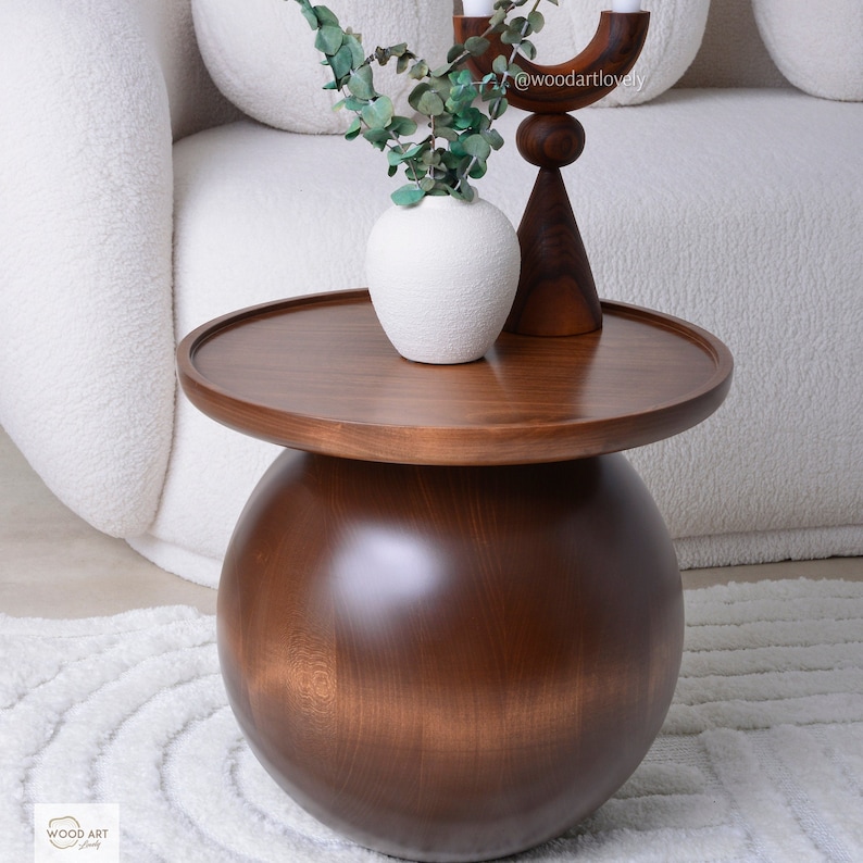 Puede incluir: Una mesa auxiliar moderna de madera marr&oacute;n oscuro con una tapa redonda y una base esf&eacute;rica. La mesa exhibe un jarr&oacute;n blanco con follaje verde y un candelabro de madera. El dise&ntilde;o es minimalista y elegante, con un di&aacute;metro de aproximadamente 60 cm.
