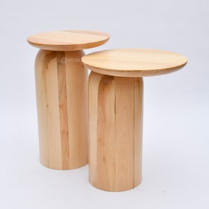 May include: Two wooden side tables with round tops and cylindrical bases. The tables are made of light wood with a natural finish. The larger table is slightly taller than the smaller table.
