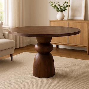 May include: A round, dark brown wooden dining table with a unique pedestal base. The base features a stacked, bulbous design. The table is set on a light beige rug in a well-lit room. The table's diameter is approximately 48 inches.