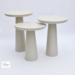 Op de afbeelding: Set van drie witte nesttafels met ronde bladen en taps toelopende poten. De tafels zijn gerangschikt in aflopende volgorde, met de grootste tafel achterin en de kleinste tafel vooraan. De tafels zijn gemaakt van hout en hebben een gladde, witte afwerking.