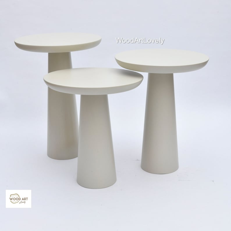 Op de afbeelding: Set van drie witte nesttafels met ronde bladen en taps toelopende poten. De tafels zijn gerangschikt in aflopende volgorde, met de grootste tafel achterin en de kleinste tafel vooraan. De tafels zijn gemaakt van hout en hebben een gladde, witte afwerking.