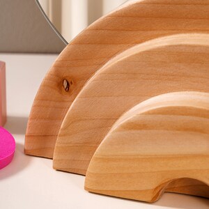 May include: A wooden mirror stand with three tiers, each shaped like a half-circle. The stand is made of light brown wood and has a natural finish.